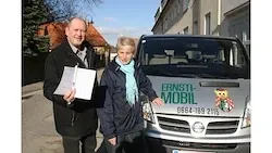 Ein Mann und eine Frau stehen neben einem Van mit dem Wort 'ERNSTI' auf der Seite. Der Mann hält ein Stück Papier, und die Frau trägt einen blauen Schal.