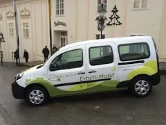 Ein weißer und grüner Van mit der Aufschrift 'Ernst-Mobil' parkt am Straßenrand vor einem weißen Gebäude, vor dem mehrere Personen stehen.