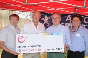 Vier Männer stehen unter einem roten Zelt und halten ein großes weißes Schild mit einem roten Logo und Text auf Deutsch.