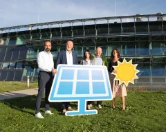 Fünf Personen stehen vor einem Solarpanel auf einem Rasenfeld, mit einem großen Gebäude im Hintergrund.