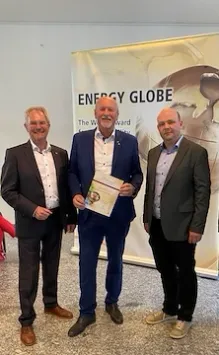 Drei Männer in Anzügen stehen vor einem Banner mit dem Text 'ENERGY GLOBE'. Der Mann in der Mitte hält ein Zertifikat.