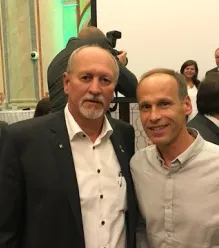 Zwei Männer stehen nebeneinander und lächeln für ein Foto. Der Mann auf der linken Seite trägt einen schwarzen Anzug und ein weißes Hemd. Der Mann auf der rechten Seite trägt ein helles Hemd. Hinter ihnen stehen mehrere Personen und unterhalten sich, und eine Kamera ist sichtbar.