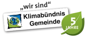 Ein weißes Schild mit einem grünen Blatt und einem Globusbild auf schwarzem Hintergrund. Der Text lautet 'Klimabündnis Gemeinde' und '5 Jahre'.