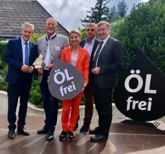 Fünf Personen stehen vor einem Gebäude mit einem Schild, das 'ÖL frei' sagt. Die Frau in der Mitte hält ein Schild, das 'ÖL frei' sagt. Der Mann auf der linken Seite hält einen Pokal.