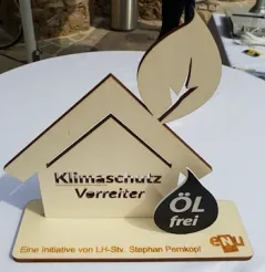 Ein Holzschild mit den Worten 'Klimaschutz Verreiter' und einem Blattdesign oben. Es hat auch ein schwarzes Öltropfensymbol mit dem Text 'ÖL frei'. Es steht auf einem weißen Tisch.