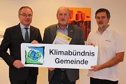Drei Männer stehen vor einer Wand und halten ein Schild, auf dem Klimabündnis Gemeinde steht. Der Mann auf der linken Seite trägt einen Anzug und eine Krawatte, der Mann in der Mitte hält ein Stück Papier und der Mann auf der rechten Seite trägt ein weißes Poloshirt.