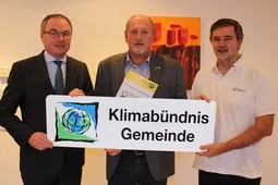 Drei Männer stehen vor einer weißen Wand und halten ein Schild mit der Aufschrift 'Klimabündnis Gemeinde'. Der Mann auf der linken Seite trägt einen schwarzen Anzug und eine Krawatte, der Mann in der Mitte hält ein Büchlein und der Mann auf der rechten Seite trägt ein weißes Hemd.