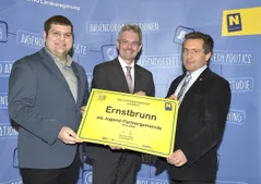 Drei Männer stehen vor einem blauen Hintergrund mit einem gelben Schild, auf dem 'Ernstbrunn als Jugend-Partnergemeinde 2016-2018' steht. Der Mann auf der linken Seite hält ein gelbes Zertifikat, und der Mann in der Mitte lächelt, während er das Zertifikat hält.