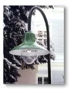 Eine grüne Straßenlampe mit einer weißen Glühbirne ist an einem schwarzen Pfahl montiert. Sie ist von einem schneebedeckten Baum und einem Gebäude mit einer rosa Wand umgeben.