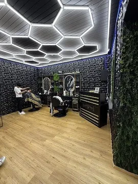 Ein moderner Friseursalon mit sechseckigem Deckendesign, Holzböden und einer grünen Wand. Ein Friseur schneidet Haare, während eine andere Person auf einem Stuhl sitzt.