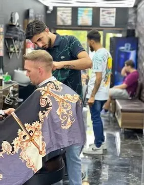 Ein Mann lässt sich in einem Barbershop die Haare schneiden. Der Friseur steht hinter ihm und schneidet ihm die Haare. Ein anderer Mann steht in der Nähe und beobachtet.