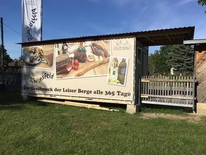 Ein kleiner Imbissstand mit einem Banner und einem Holzzaun. Das Banner zeigt Bilder von Essen und Getränken und der Text lautet 'Der Geschmack der Leiser Berge alle 365 Tage'.