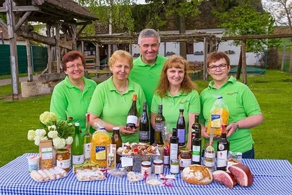 Eine Gruppe von Menschen in grünen Hemden steht hinter einem Tisch voller verschiedener Lebensmittel, darunter Brot, Käse und Weinflaschen.