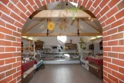 Ein Innenraum einer Bäckerei mit Ziegelwänden, Holzbalken und einem gefliesten Boden. Es sind verschiedene Lebensmittel ausgestellt, darunter Brot, Gebäck und andere Backwaren.
