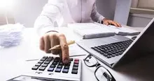 Eine Person arbeitet an einem Schreibtisch mit einem Taschenrechner, Laptop und Papieren.