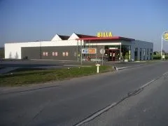 Ein großes Gebäude mit einem roten Dach und dem Wort 'BILLA' in Gelb obenauf. Das Gebäude hat mehrere Fenster und Türen. Es gibt einen gelben Feuerhydranten an der Straßenseite.