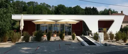 Ein modernes weißes Gebäude mit einem geschwungenen Dach und Glasfenstern. Vor dem Eingang stehen zwei große Sonnenschirme.