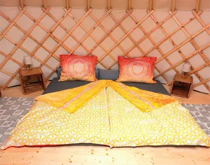 Ein gemütliches Bett in einem Jurten mit einer gelben Decke, zwei roten Kissen und zwei kleinen hölzernen Nachttischen mit Lampen.