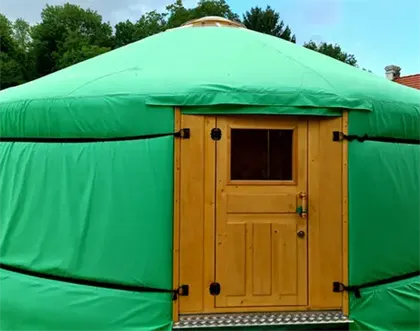 Ein grünes Jurtenzelt mit einer Holztür und einem Fenster steht in einem Grasbereich mit Bäumen im Hintergrund.
