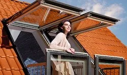 Eine Frau steht auf einem Balkon mit offenen Fenstern und einem Dachfenster auf einem roten Dach.