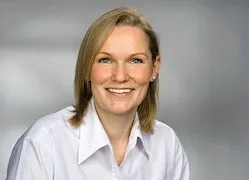 Eine Frau mit kurzen blonden Haaren lächelt und schaut in die Kamera. Sie trägt ein weißes Hemd.