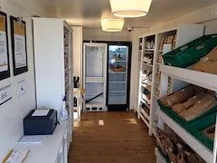 Ein Innenansicht eines kleinen Ladens mit Holzböden und weißen Wänden. Auf beiden Seiten befinden sich Regale mit verschiedenen Artikeln, und in der Mitte steht ein Kühlschrank. Ein schwarzer Drucker steht auf der linken Seite, und einige Papiere liegen auf dem Schreibtisch.