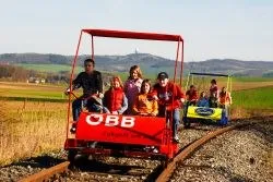 Eine Gruppe von Menschen fährt auf einem roten und gelben Zug auf einer Eisenbahnstrecke in einem ländlichen Gebiet.