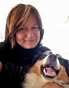 Eine Frau mit kurzen blonden Haaren lächelt und hält einen Hund. Der Hund ist ein Berner Sennenhund mit braunem, weißem und schwarzem Fell. Die Frau trägt ein schwarzes Shirt.