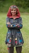 Eine ältere Frau mit einem Lächeln im Gesicht steht in einem Grasfeld mit verschränkten Armen. Sie trägt einen bunten Mantel mit Kapuze und eine Halskette.
