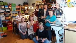 Eine Gruppe von Frauen posiert für ein Foto in einem Raum voller verschiedener Gegenstände, darunter Lagerboxen, eine Nähmaschine und ein Schreibtisch.