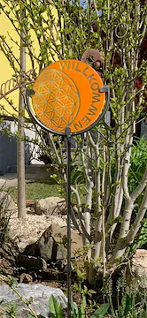 Ein orangefarbenes kreisförmiges Schild mit einem Flower-of-Life-Design steht in einem Garten.