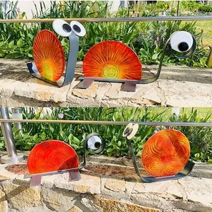 Eine Metallskulptur einer Schnecke mit großen Augen und einem orangefarbenen Gehäuse auf einer Steinmauer.