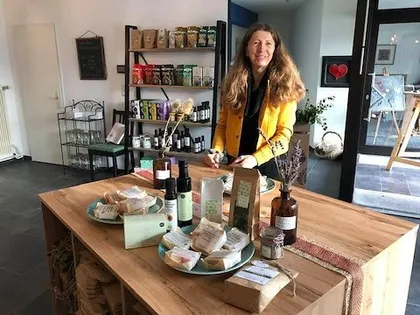 Eine Frau steht hinter einem Holztresen in einem Geschäft, lächelt und ist von verschiedenen Produkten wie Seifen, Ölen und Kräutern umgeben.