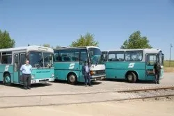 Drei türkisfarbene Busse sind auf einer Betonfläche geparkt, wobei zwei Personen vor jedem Bus stehen. Die Busse haben den Buchstaben 'S' auf ihren Fronten.
