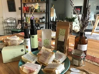 Eine sorgfältig arrangierte Einzelhandelspräsentation von natürlichen Hautpflege- und Wellnessprodukten auf einem Holztisch. Die Kollektion umfasst handwerkliche Seifen in verschiedenen Farben und Texturen, Glasflaschen mit Ölen oder Seren, Kraftpapierverpakungen, getrocknete Blumen in einer braunen Vase und Dekorationselemente. Im Hintergrund sind Regale mit zusätzlichen Produkten und Dekoration sichtbar.