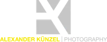 Das Logo für Alexander Kunzel Fotografie zeigt ein fettgedrucktes, weißes 'X' auf einem grauen Hintergrund. Der Name 'Alexander Kunzel' ist in einer modernen, serifenlosen Schrift unter dem 'X' geschrieben, und das Wort 'Fotografie' ist in einer kleineren, kursiven Schrift unter dem Namen geschrieben.