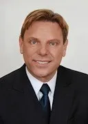 Ein Mann in einem schwarzen Anzug und blauer Krawatte lächelt und schaut direkt in die Kamera.
