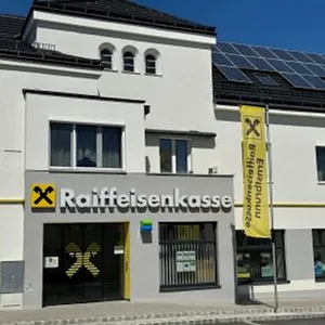 Ein weißes Gebäude mit einem schwarzen Dach und Solarpanelen auf der Oberseite. Das Gebäude hat ein Schild, das 'Raiffeisenkasse' mit einer gelben Fahne mit dem Text 'Erneuerbare Energien' liest.