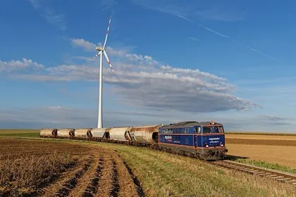 Ein Zug fährt auf der Bahnstrecke in einem Feld. Ein Windrad steht hinter dem Zug.