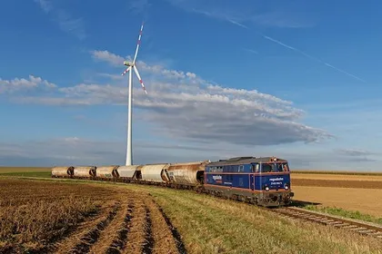 Ein blauer Zug mit mehreren Güterwaggons fährt auf einer Bahnstrecke in einem ländlichen Gebiet. Im Hintergrund steht eine große Windkraftanlage.