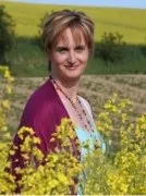 Eine Frau mit kurzen Haaren steht in einem Feld gelber Blumen, lächelt und schaut in die Kamera. Sie trägt einen rosa Cardigan und eine Halskette.