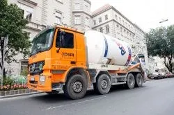 Ein orangefarbener Zementlastwagen fährt eine Stadtstraße hinunter, mit Gebäuden und Bäumen im Hintergrund.