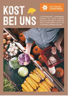 Ein Mann auf einem Marktstand wählt Gemüse aus. Er hat einen Korb in der Hand und ist von einer Vielzahl von Gemüsesorten umgeben, darunter Kürbisse, Kohl und Mais.
