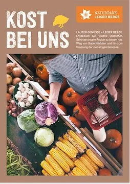 Ein Mann holt Gemüse an einem Marktstand. Der Stand ist mit einer Vielzahl von Gemüsesorten wie Kürbissen, Mais, Kohl, Zwiebeln und Bohnen gefüllt. Der Stand hat ein Schild, das 'KOST BEI UNS' besagt.