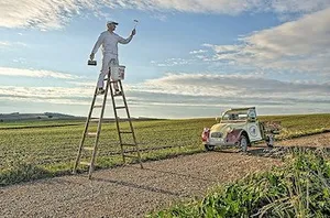 Ein Mann steht auf einer Leiter in einem Feld, hält einen Pinsel und eine Dose Farbe. Er ist in einem weißen Anzug und Hut gekleidet.