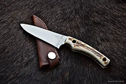 Ein scharfes Messer mit weißem Griff und brauner Lederscheide liegt auf einer braunen Pelzoberfläche.