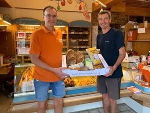 Zwei Männer in einem Bäckereiladen, einer trägt ein orangefarbenes Poloshirt und der andere ein blaues T-Shirt. Sie halten eine Schachtel mit Brot und Gebäck.