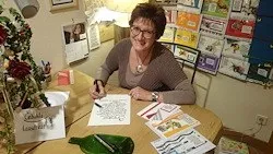 Eine Frau mit Brille und Halskette sitzt an einem Tisch mit Papieren und einem Stift, lächelt und schaut in die Kamera.