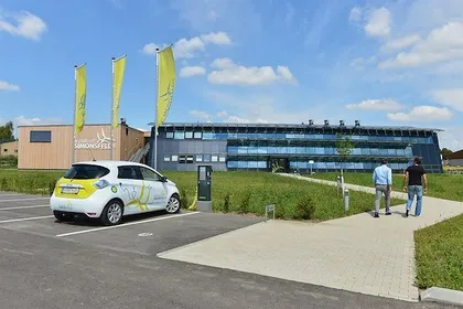 Ein weißer und gelber Elektroauto ist vor einem modernen Gebäude mit Glasfenstern geparkt. Zwei Männer gehen auf einem gepflasterten Weg vor dem Gebäude.