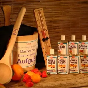 Eine Saunaausstattungskollektion auf einer Holzoberfläche mit hölzernen Kellen und Bürsten, einem schwarzen Saunaeimer mit der Aufschrift 'Aufguss', dekorativen Steinen in Orange- und Rottönen sowie mehreren Flaschen Saunaessenz in einer Reihe. Die warme Beleuchtung und der Holzhintergrund schaffen eine einladende Spa-Atmosphäre.